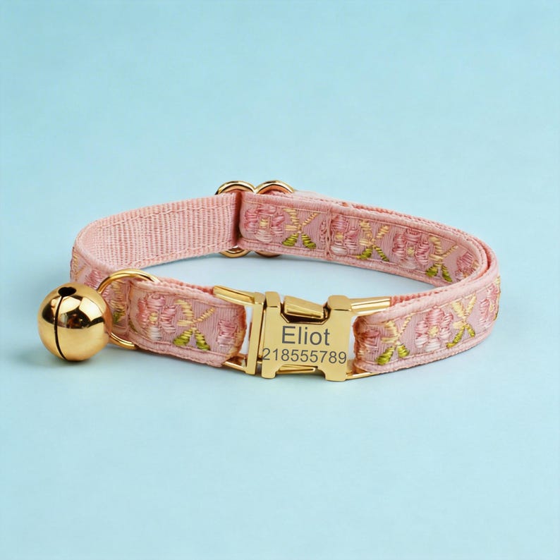 May include: A light pink pet collar featuring an embroidered floral pattern in shades of pink and green. The collar includes a gold-toned metal buckle engraved with the name Eliot and the number 218555789, along with a matching gold-toned bell.