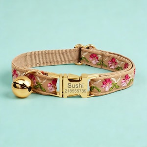 May include: A beige pet collar featuring an embroidered floral pattern in pink and green. The collar includes a gold-toned metal buckle engraved with the name Sushi and the number 218555789, along with a matching gold-toned jingle bell attached.