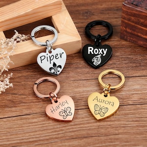 May include: Heart-shaped keychains in silver, black, rose gold, and gold. Each keychain has a name engraved on it: Piper, Roxy, Harley, and Aurora. The keychains also feature decorative elements like a fleur-de-lis and a butterfly.