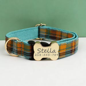 May include: A pet collar featuring a teal and orange plaid pattern with a gold-toned metal D-ring and a bone-shaped gold-toned buckle. The buckle is engraved with the name Stella and the phone number 218-555-789 in a dark, legible font.
