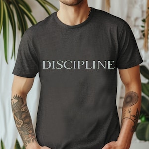 May include: Dark gray t-shirt with the word "DISCIPLINE" printed in a light gray color across the chest. The shirt is made of a soft material and has short sleeves. The model has tattoos on their arms.