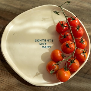 Puede incluir: Un plato de cerámica de color crema, de forma irregular, con un racimo de tomates cherry rojos maduros en una rama. El plato tiene el texto "CONTENTS MAY VARY" en azul. El plato está sobre una superficie de madera.