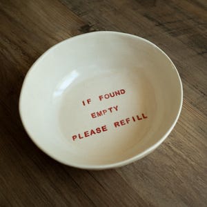 Puede incluir: Un cuenco redondo de cerámica de color blanco roto con un acabado liso. El interior del cuenco presenta el texto "IF FOUND EMPTY PLEASE REFILL" impreso en una fuente sans-serif roja, sencilla y negrita, centrada en la base.