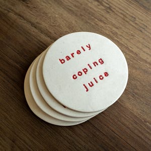 Pode incluir: Uma pilha de bases redondas brancas com as palavras "barely coping juice" em letras vermelhas. As bases estão sobre uma superfície de madeira escura.