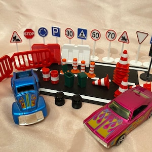 May include: A set of toy road signs, traffic cones, barriers, and two toy cars. The signs display speed limits, stop signs, and other road symbols. The cars are blue and pink with flame designs.