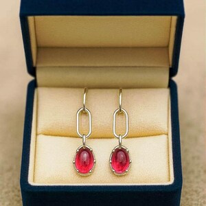 Puede incluir: Un par de pendientes colgantes plateados con piedras preciosas rojas ovaladas. Los pendientes se muestran en una caja de joyería de terciopelo azul marino abierta.