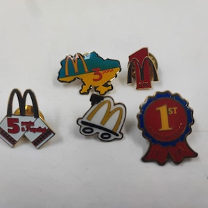 May include: A collection of enamel pins featuring the McDonald's logo and related designs. Includes a pin shaped like the Ukrainian map with the McDonald's logo, a "1st" place ribbon pin, and pins with the McDonald's arches.