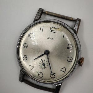 May include: Vintage wristwatch with a silver face, black hands, and black numerals. The watch has a worn, silver-colored metal case and a gold-colored crown. The watch face has the word "Зим" printed on it.