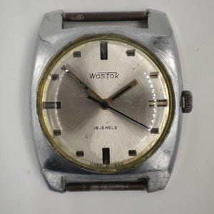 Puede incluir: Reloj de pulsera Wostok vintage plateado con esfera redonda y clara. La esfera presenta agujas y marcadores negros, con la marca "Wostok" y "18 JEWELS" impresas. La caja es rectangular y tiene una corona en el lateral.