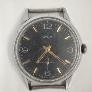Peut inclure: Une montre-bracelet vintage avec un cadran noir, des aiguilles et des index dorés, et un boîtier en métal argenté. Le cadran affiche les chiffres 12, 3, 6 et 9, avec le texte "Made in USSR" en bas. La montre a une petite trotteuse.
