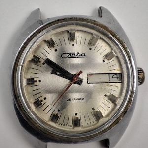 May include: A vintage silver-toned wristwatch with a light silver face. The watch face features black hour markers, black hands, and a date display at the 3 o'clock position. The watch has the word "Слава" on the face.