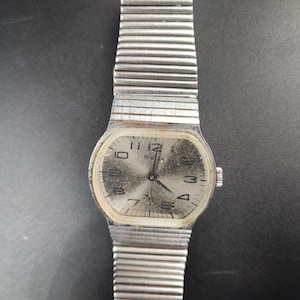 May include: A vintage silver wristwatch with a rectangular face and a ribbed metal band. The watch face displays the time with black numerals and hands. The watch has the word "ПОБЕДА" on the face.
