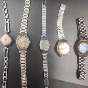 May include: Five vintage wristwatches with silver-toned metal bands and watch faces. The watch faces vary in shape and color, including square, round, and rectangular designs. The watches are displayed on a dark surface.