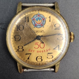 Puede incluir: Reloj de pulsera vintage dorado con esfera redonda. La esfera del reloj presenta números negros, manecillas negras y el texto "50 ЛЕТ ПОБЕДЫ". Un emblema de estrella está en la parte superior. El texto "СДЕЛАНО В РОССИИ" está en la parte inferior.