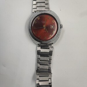 Peut inclure: Une montre-bracelet vintage avec un bracelet et un boîtier en métal argenté. Le cadran de la montre est de couleur rouge foncé avec des aiguilles et des marqueurs dorés. Une petite fenêtre de date est visible à 3 heures. La montre a une lunette cannelée.