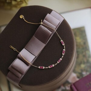 May include: A delicate gold bracelet with rectangular ruby accents and small white stones. The bracelet is displayed on a brown velvet box with a satin ribbon. The jewelry is elegant and suitable for special occasions.