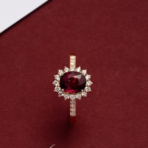 May include: A gold ring featuring a large, oval-cut, deep red gemstone surrounded by a halo of smaller, clear gemstones. The ring's band is also adorned with small, clear gemstones. The ring is set against a dark red background.