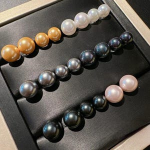 May include: Three hair clips featuring rows of spherical pearls. The top clip has white pearls, the middle clip has gold-toned pearls, and the bottom clip features a gradient of dark grey to light pink pearls, all arranged in a linear fashion.