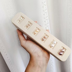 Puede incluir: Exhibición de pendientes de perlas en varios colores, incluyendo blanco, rosa y crema. Los pendientes se presentan en un soporte rectangular beige. El soporte está siendo sostenido por una persona.