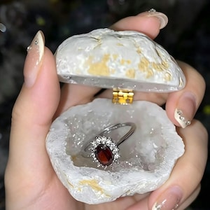 May include: A silver ring with a dark red oval gemstone, encircled by smaller clear stones, presented in an open, white geode-shaped jewellery box. The geode box has a gold hinge. The ring is the focal point.