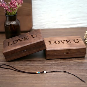 Puede incluir: Dos cajas rectangulares de madera con el texto grabado "LOVE U" en las tapas. Las cajas son de madera marrón oscuro y tienen un diseño sencillo y elegante. Un cordón marrón con cuentas decorativas se encuentra frente a las cajas. Un jarrón con flores rosas está en el fondo.
