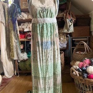 May include: A long, flowing white dress with a green and blue floral print. The dress has a white sash around the waist and a beaded trim at the hem.