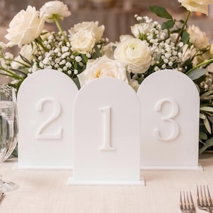 May include: Elegant wedding table setting with white roses, greenery, and lit candles. Features white table number cards with the numbers 1, 2, and 3. Includes plates, silverware, and wine glasses, creating a sophisticated ambiance.