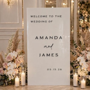Può includere: Un cartello nuziale bianco con testo nero: "WELCOME TO THE WEDDING OF AMANDA and JAMES 05.15.26". Composizioni floreali con fiori bianchi e rosa, erba della pampa e candele fiancheggiano il cartello.
