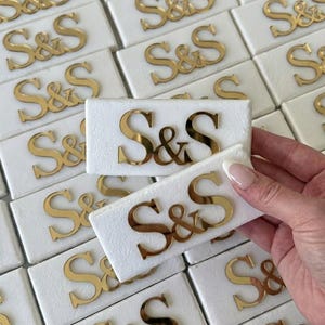 May include: Close-up of multiple white rectangular objects with gold-colored mirrored letters spelling "S&S". One object is held by a hand. The objects are arranged closely together, creating a pattern. The letters are in a stylized font.