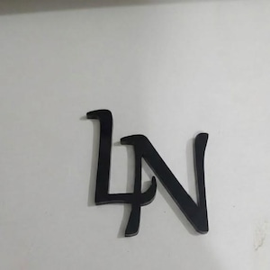 Pode incluir: Letras pretas de metal 'LN' em uma fonte estilizada, repousando sobre uma superfície branca. As letras estão conectadas e têm uma aparência moderna e elegante. A imagem é bem iluminada, destacando a textura suave das letras.
