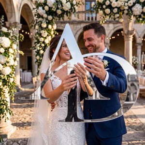 May include: A large, reflective, silver-toned acrylic sign featuring the capital letters A and D intertwined in a stylized monogram. The surface of the sign acts as a mirror, reflecting the image of a person in a white lace wedding dress.