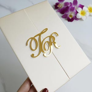 Può includere: Invito di nozze color avorio con monogramma dorato delle lettere "JR". L'invito è un design a due ante con una superficie strutturata. Fiori viola e bianchi sullo sfondo.