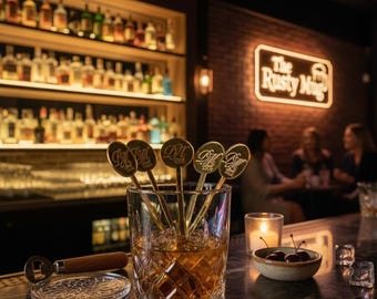 May include: A rocks glass filled with a brown liquid and gold bar spoons. The spoons have a circular design with the letters "DM" on them. A bottle opener and coaster are on the bar. The background shows a bar with bottles and a neon sign that says "The Rusty Mug".