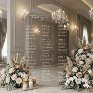 May include: An arched, clear acrylic wedding seating chart with white text reading Find Your Seat, listing guests by table numbers from 1 to 12. The sign is displayed on a marble surface, flanked by two large floral arrangements of white roses.