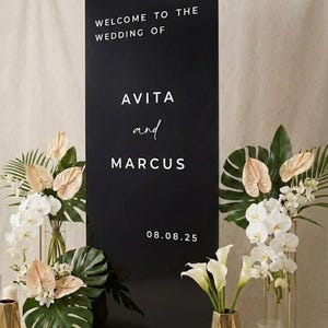 Könnte beinhalten: Schwarzes Hochzeitsschild mit weißem Text: "WELCOME TO THE WEDDING OF AVITA and MARCUS 08.08.25". Das Schild ist von Blumengestecken mit weißen und pfirsichfarbenen Blumen umgeben.