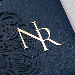 May include: A close-up of a navy blue invitation with gold foil lettering and intricate laser-cut detailing. The gold letters "NR" are prominently displayed, set against a shimmering background. The design features a delicate, ornamental border.