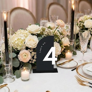 May include: A black table number 4 with white text, set on a white tablecloth. The table is decorated with white flowers, candles, and gold-rimmed plates, creating an elegant wedding or event setting.