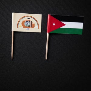 May include: Two small flags on wooden sticks. One flag features a rainbow over a building and a landscape. The other flag displays the flag of Jordan, with red, black, white, and green stripes and a white star.