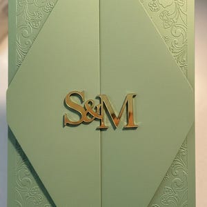 May include: A sage green rectangular invitation card featuring a gatefold design with a diamond-shaped center panel. The card is embossed with floral patterns and displays the gold metallic letters S&M centered on the front closure.