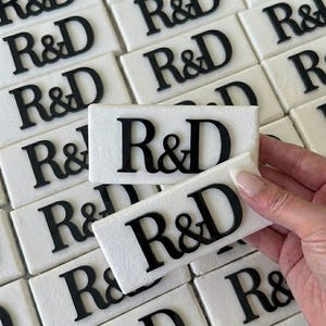 May include: Rectangular white tiles with the black letters "R&D" are arranged in a grid. One tile is held in a hand. The tiles have a textured surface. The letters are in a bold, serif font.