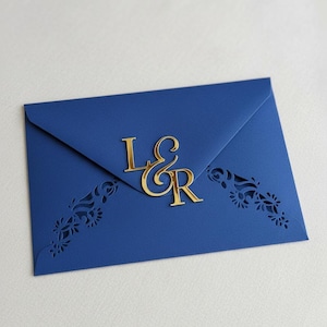 May include: A navy blue envelope with gold-colored initials "L&R" on the flap. The envelope has decorative floral cutouts on the edges. The background is a plain white surface.