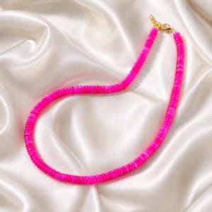 May include: A vibrant hot pink beaded necklace featuring small, flat, circular disc-shaped beads strung together. The necklace is finished with a gold-toned metal lobster claw clasp and a short extender chain at the end for adjustable length.
