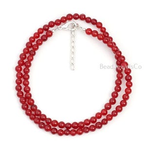 Peut inclure: Un collier à double rangée composé de petites perles rondes rouge foncé. Le collier est doté d'un fermoir et d'une chaîne d'extension argentés. Le nom de la marque "Bead Jewels Co" est imprimé en blanc sur le côté droit.