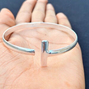 May include: A silver-colored cuff bracelet with a T-shaped bar in the center. The bracelet is open-ended and appears to be made of polished metal. The jewelry is shown on a person's hand.