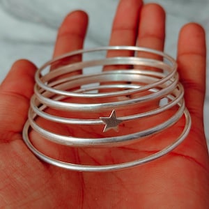 May include: A stack of silver bangles, one featuring a star charm. The bracelets are round and smooth, reflecting light. The jewelry is held in a hand, showcasing the design and texture of the metal.