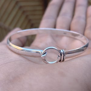 May include: A silver bangle bracelet with a hook and eye clasp. The bracelet is round and has a smooth, polished finish. The clasp is a circular design with a hook closure. The bracelet is shown on a hand.
