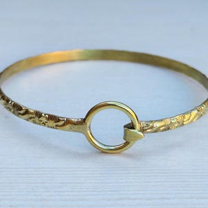 Op de afbeelding: Een goudkleurige armband met een gegraveerd bloemmotief. De armband heeft een cirkelvormige sluiting en is ongeveer 6,35 cm in diameter. Het ontwerp is eenvoudig en elegant, geschikt voor dagelijks gebruik.