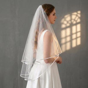 Peut inclure: Un voile de mariée blanc à deux niveaux orné d'un délicat ruban de satin sur les bords. Le tissu en tulle transparent tombe doucement sur les épaules, créant un effet superposé qui complète la robe blanche sans manches portée en dessous.