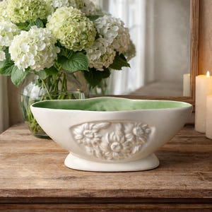 Puede incluir: Un cuenco decorativo de cerámica blanca con interior verde y diseño floral en relieve. El cuenco está sobre una superficie de madera, con un jarrón de flores blancas al fondo. También se ven dos velas encendidas y un espejo.