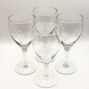 May include: Four clear wine glasses with etched grape cluster designs. The glasses have a classic shape with a round base, a slender stem, and a bowl that tapers towards the top. The etched design adds a decorative touch to the glassware.